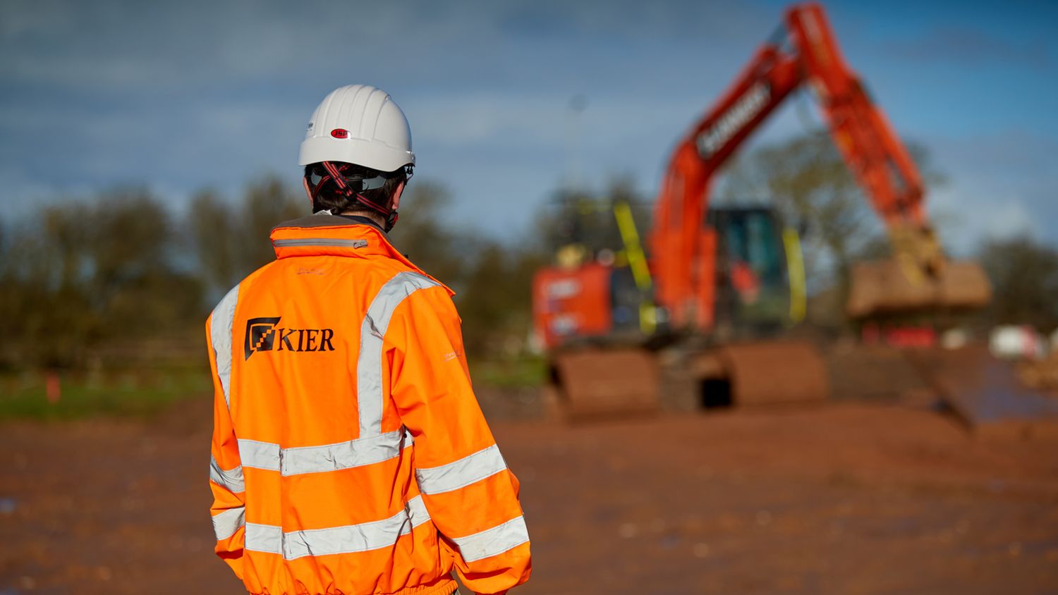 A Kier worker in an orange hi-vis jacket and white hard hat stands on a site with a red excavator in the background.