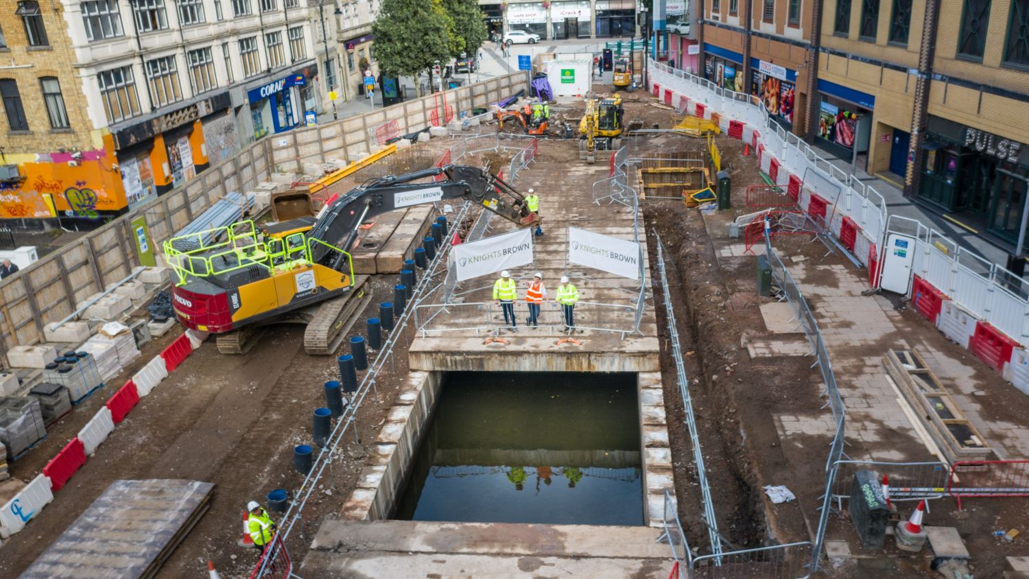 Aerial shot of the uncovered dock feeder canal in Churchill Way, Cardiff. Three individuals in hi-vis stand close to the open canal with an excavator's arm arched behind them (Image courtesy of Atkins)