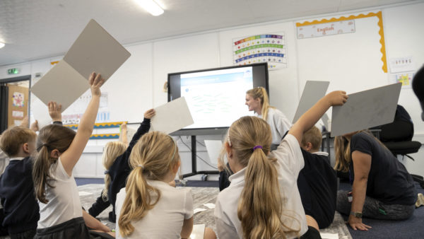 Children putting their hands up in class as a female teacher leaders a lesson (Image courtesy of Developing Experts)