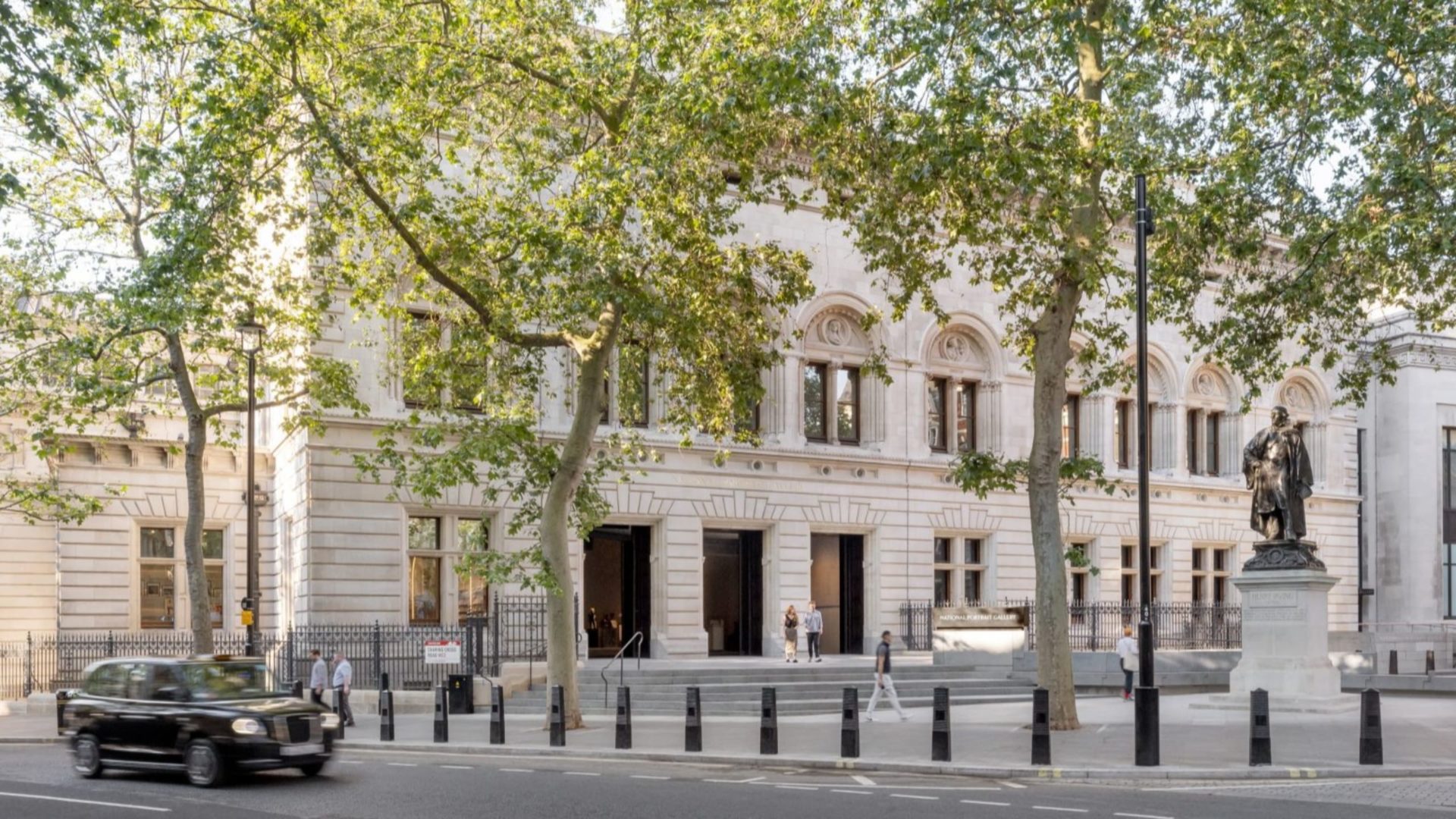 Front of an old white stone building with a black cab on the road and a statue and trees in the background.