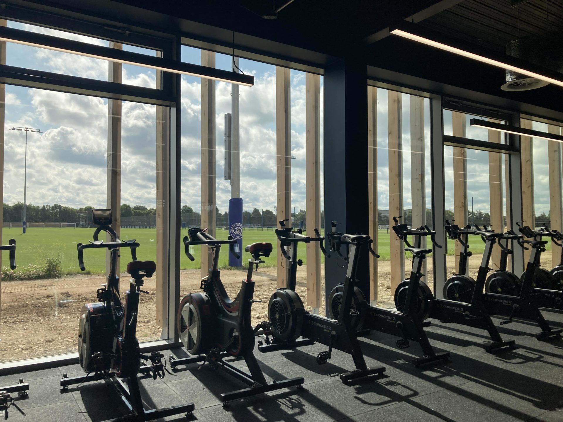 Buro Happold QPR - Exercise bikes aligned in front of a window. There's a path and green grass behind the window.