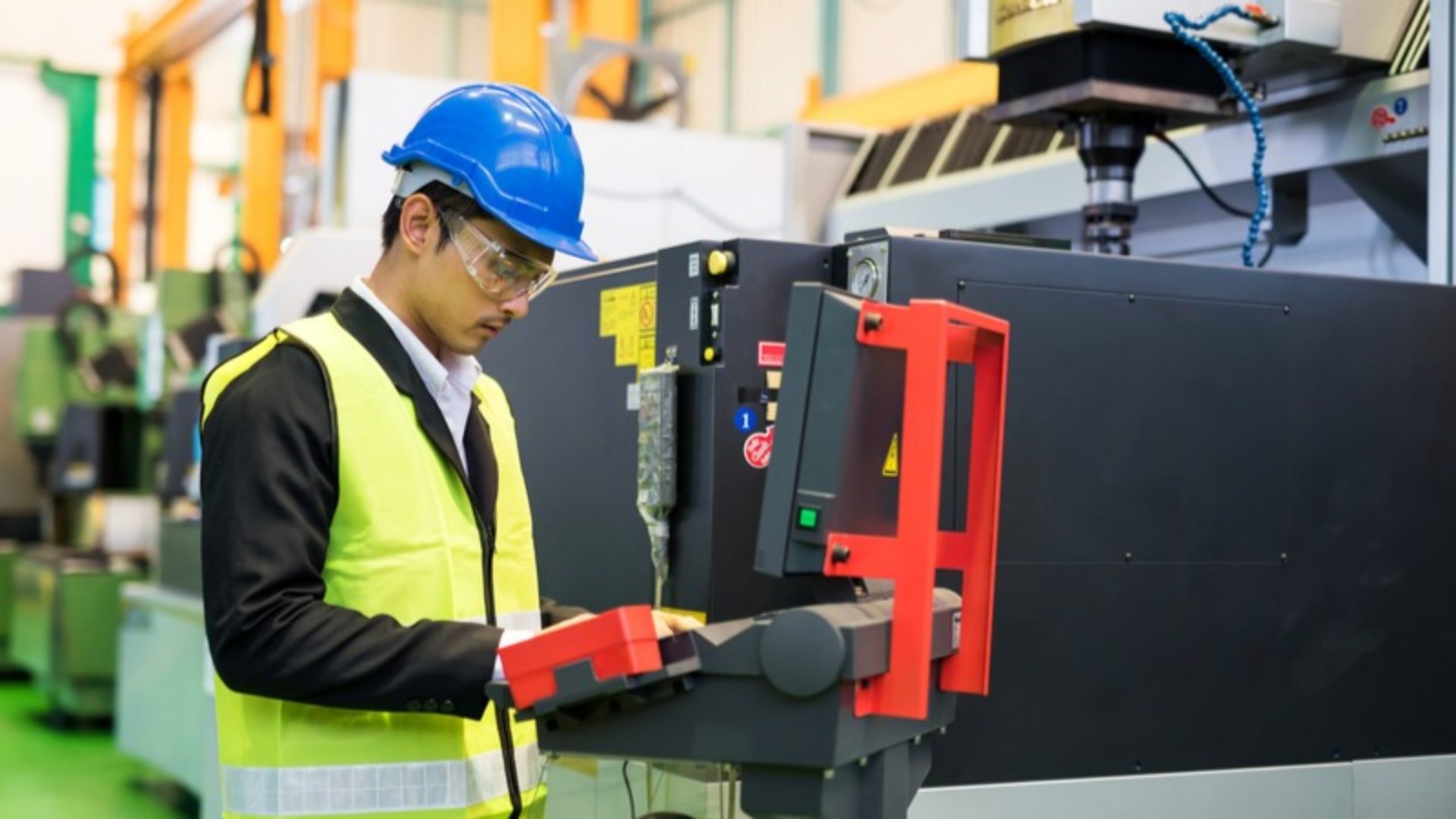 A waist-up man with hardhat and protection glasses using a computer in a factory.