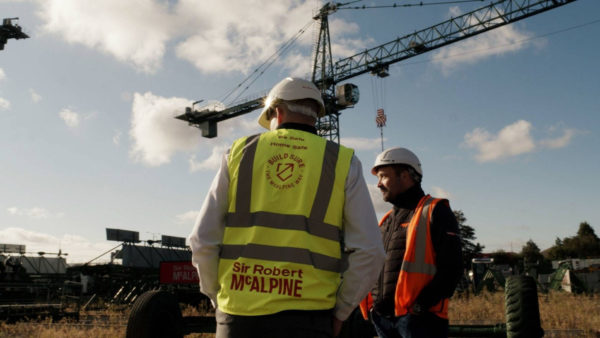 Two construction workers with hard hats and high-vis jackets and a crane in the background.