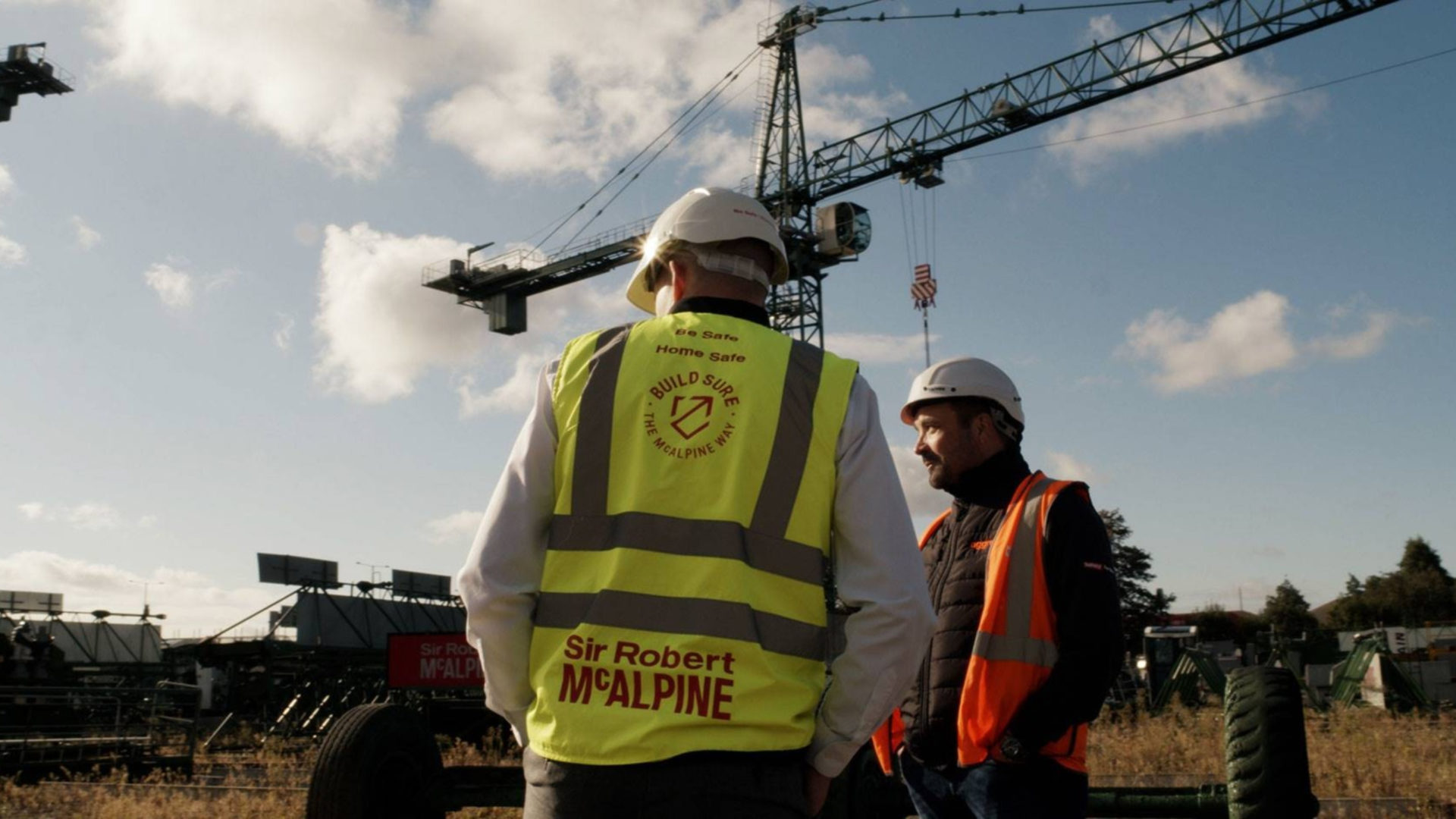 Two construction workers with hard hats and high-vis jackets and a crane in the background.