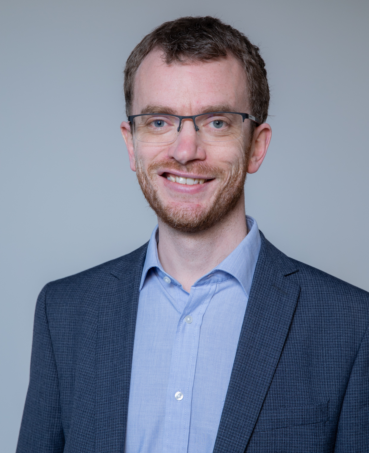 Mugshot of a man with glasses and blue shirt and jacket smiling at the camera - he is the author of this piece on the impact of Trump's tariffs in construction, his name is Chris Kirby-Turner, a partner at Thomson Snell & Passmore