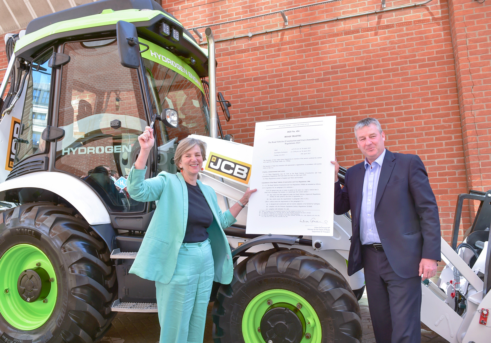 A woman and a man holding a big paper in front of a green digger - the government has changed the law to give hydrogen diggers the green light to be driven on UK roads.