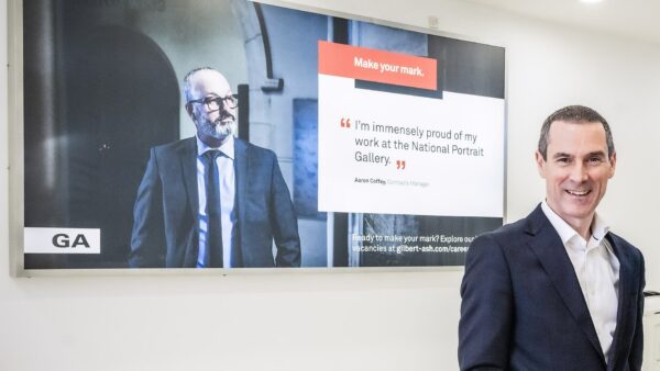 A man in front of a poster that has another man and a quote - Gilbert-Ash has transitioned to an employee owned trust.