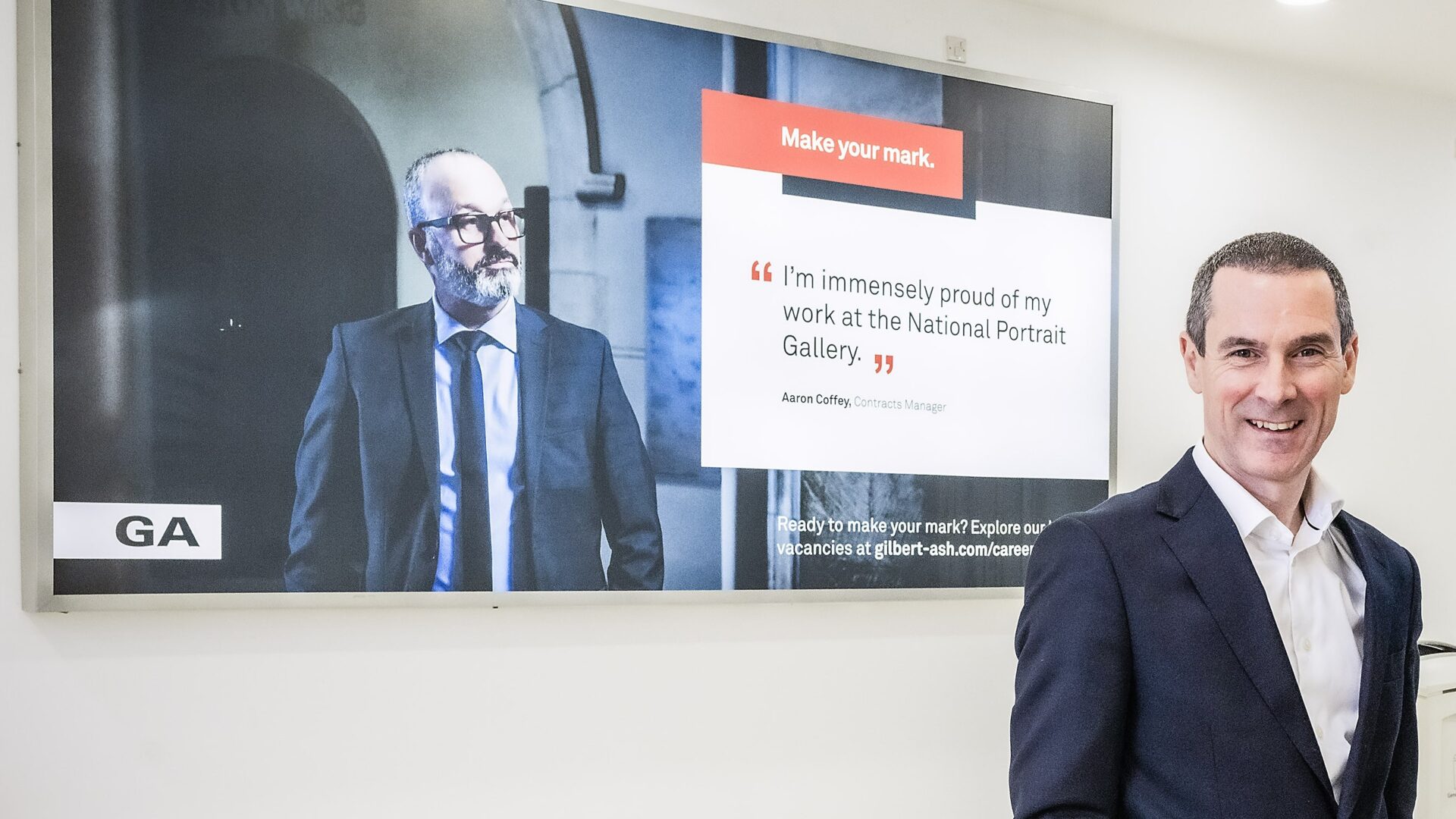 A man in front of a poster that has another man and a quote - Gilbert-Ash has transitioned to an employee owned trust.