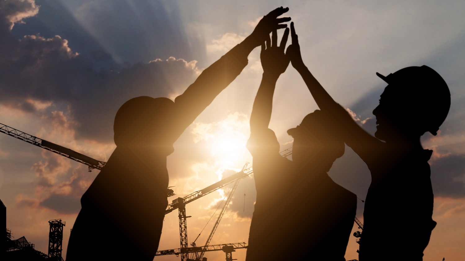 Silhouette of three workers wearing hard hats against a sunset background with cranes