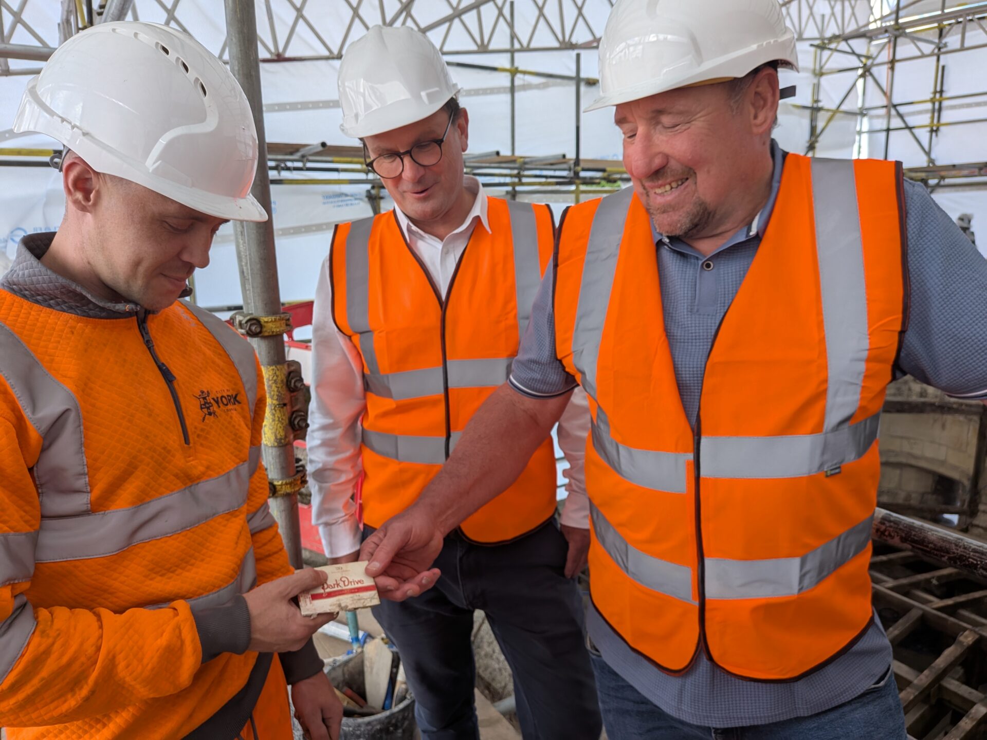 Three men of different ages with hard hats and orange high-vis jackets looking at an old cigarette box