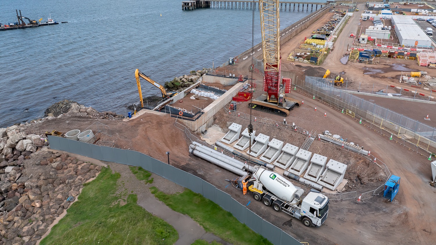 A construction site next to the sea with. There's a big white concrete mixer truck and a yellow crane.