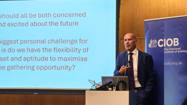 A man talking behind a lectern with a screen behind him - Mark Farmer delivering the 2025 CIOB Sir James Wates Lecture